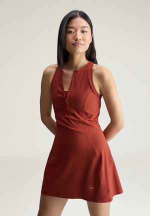 Femme aux longs cheveux foncés portant une robe de sport sans manches couleur rouille avec une fermeture éclair à l'avant et des perforations sur les côtés, debout les mains derrière le dos.