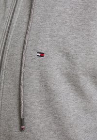 Graue Zip-Hoodie aus weichem Stoff, mit einem kleinen gestickten Logo in Schwarz, Weiß und Rot. Enthält geflochtene Kordeln.