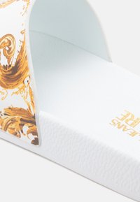 Weiße Pantoletten mit gummierter Profilsohle. Der Riemen ist mit einem dekorativen gold-braunen Muster versehen, die Innenseite ist glatt.