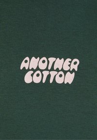 Cămașă din bumbac verde închis, cu un grafic de text alb mare care spune "ANOTHER COTTON" într-un font rotund și jucăuș.