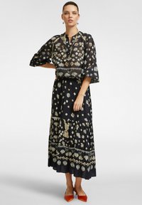 Lungo abito nero con motivi floreali in crema e oro. Blusa trasparente con maniche a campana, gonnellina a più strati e tacchi arancioni.