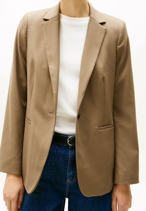 Person iført en beige blazer over en hvid skjorte med blå jeans og sort bælte, stående mod en ensfarvet baggrund.