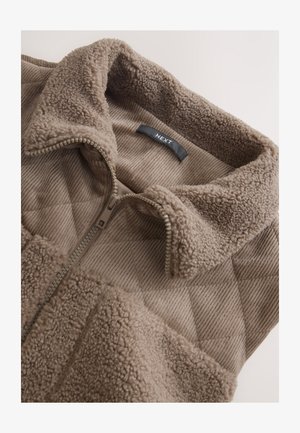 Veste à fermeture éclair en polaire marron avec un col texturé, un motif à rayures diagonales, et une étiquette noire à l'intérieur. Dotée d'une fermeture éclair solide et d'un matériau doux.