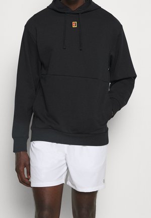 Homme portant un sweat à capuche noir avec un petit logo rouge et jaune sur la poitrine et un short de sport blanc, debout avec une main dans la poche.