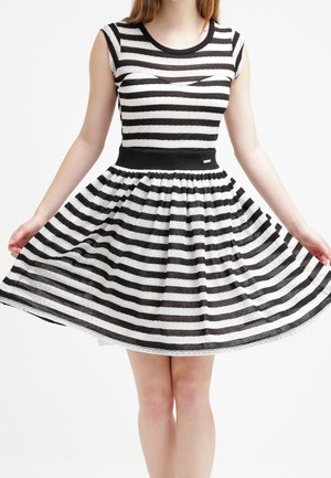 Frau trägt ein schwarz-weiß horizontal gestreiftes ärmelloses Kleid und hält den Rock auf beiden Seiten seitlich ausgestreckt.