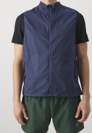 Homme portant une veste zippée bleu marine sur un t-shirt noir à manches courtes et un short de sport vert foncé, debout devant un fond neutre.