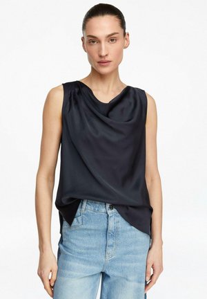 Kobieta w czarnym, bezrękawnym satynowym topie z drapowaniem, włożonym w jasno-niebieskie, wysokim stanem jeansy, stojąca na białym tle.
