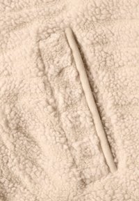 Gros plan d’un tissu polaire sherpa beige avec une poche verticale dotée d’une ouverture bordée de tissu.