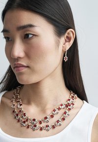 Náhrdelník s vrstveným designem, zdobený červenými a fialovými kameny zasazenými do zlatě tónovaného kovu, doplněný ovislými náušnicemi se shodnými akcenty červeného kamene.