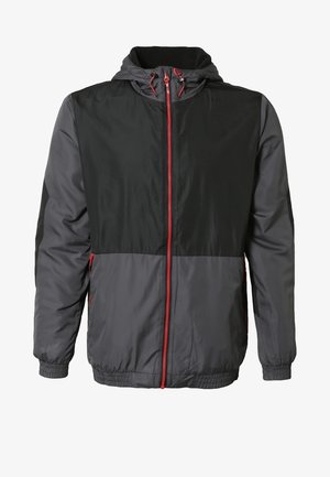 Veste d'homme légère, noire et grise avec des accents rouges, associée à un pantalon cargo beige et des chaussures de randonnée grises. Texture lisse, design à capuche.