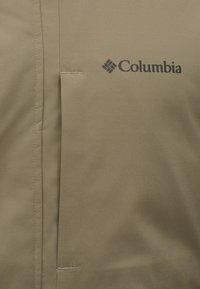 Μπεζ υφασμάτινο πάνελ με ραμμένη ραφή και μαύρο λογότυπο "Columbia" τυπωμένο στην επάνω δεξιά πλευρά.