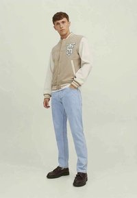 Casaco varsity bege e creme com um logo gráfico, punhos canelados em bege, combinado com jeans azul claro e sapatos castanhos escuros.