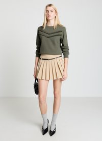 Pull vert olive avec détails en chevrons à franges, mini-jupe plissée beige, chaussettes grises, et escarpins noirs à bout pointu. Tient un petit sac noir.