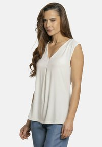 Blusa blanca sin mangas con diseño de cuello en V, tela fluida, pliegue sutil en la parte delantera y un corte holgado. Combinada con denim azul claro.