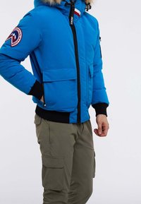 Man med blå vinterjacka med pälsluva och gröna cargobyxor, med handen i jackfickan, stående mot vit bakgrund.