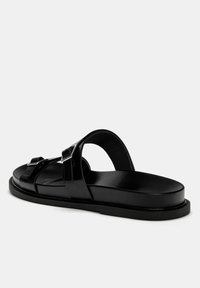 Sandale en cuir noir avec un dos ouvert, présentant deux sangles réglables et des boucles en acier inoxydable, semelle plate et texture lisse.