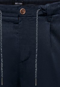 Pantalon marine foncé avec cordons tressés, fermeture à bouton marron et étiquette de marque "Only & Sons" à l'intérieur de la ceinture.