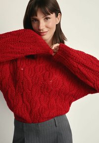 Czerwony, gruby sweter dzianinowy z wzorem warkocza, o luźnym kroju i szerokich rękawach. Noszony z szarymi spodniami w prążki, ukazującymi fakturowany wzór.