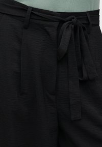 Μαύροι παντελόνια με υφή και υφασμάτινη ζώνη δεμένη σε φιόγκο στη μέση, φορεμένα με πράσινο τοπ μέσα από την παντελόνα.