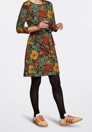 Kleid mit Blumenmuster in Grün, Orange und Gelb, mit Taillengürtel und Dreiviertelärmeln. Kombiniert mit hellbraunen und roten Turnschuhen.