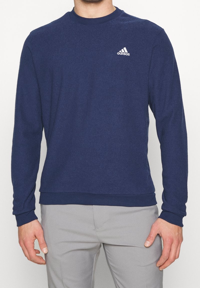 Homme portant un sweat-shirt Adidas à manches longues bleu marine et un pantalon gris clair, debout les bras détendus le long du corps.