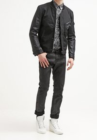 Giacca bomber in pelle nera sopra una camicia a fantasia, abbinata a jeans scuri e sneakers bianche alte. Caratteristiche principali: vestibilità su misura e design elegante.
