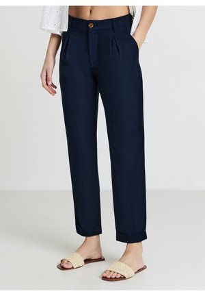 Femme portant un pantalon bleu marine taille haute avec plis sur le devant et revers roulés, associé à des sandales plates tissées beiges.
