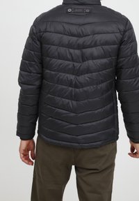Veste matelassée noire avec rembourrage matelassé horizontal, col montant et finition texturée. Comprend une étiquette de marque et des poignets élastiques.