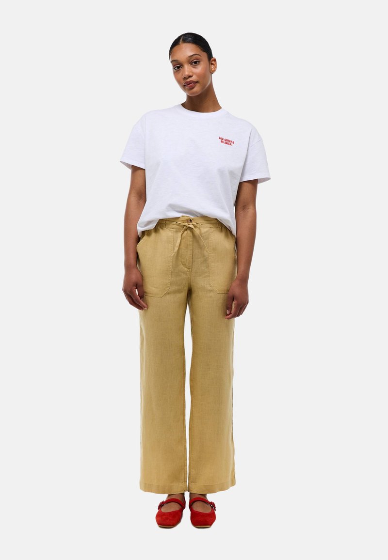 T-shirt en coton blanc avec texte rouge, associé à un pantalon en lin beige clair avec taille à cordon et deux poches avant. Chaussures rouges avec des nœuds.