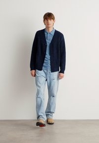 Cárdigan de punto navy sobre una camisa de cuello azul claro, combinado con jeans desgastados azul claro y zapatos beige con acentos negros.