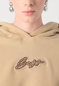 Beige hettegenser med en teksturert, brodert logo med brune og gull perler. Stoffet har en myk, glatt overflate.
