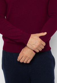 Bordeauxrode gebreide trui met lange mouwen en geribde manchetten, gecombineerd met donkerblauwe broek. Close-up van handen die elkaar voor de taille kruisen.