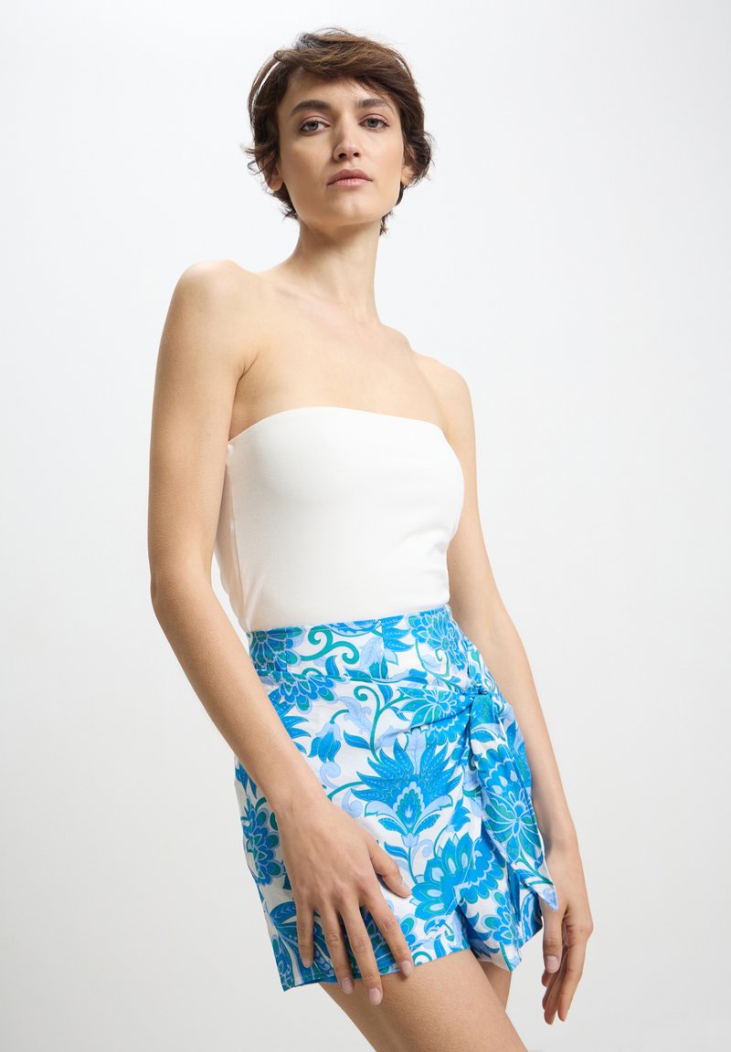 Donna che indossa un top bianco senza spalline e una gonna avvolgente blu con motivi floreali, in piedi di fronte a uno sfondo bianco semplice.