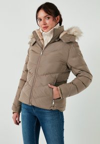 Giubbotto imbottito color taupe con cappuccio foderato e rifiniture in pelliccia sintetica. Presenta cuciture orizzontali e tasche frontali con zip. Indossato con un dolcevita color crema.