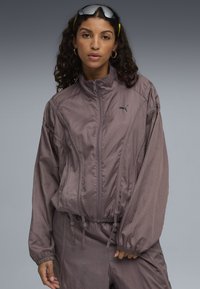 Puma zip-up jas in gedempt paars, lichtgewicht stof, elastische manchetten, trekkoord bij de zoom, minimalistisch ontwerp, met een klein logo op de borst.