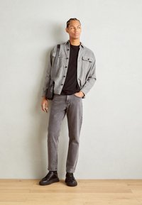 Chemise grise boutonnée portée sur un t-shirt noir, associée à un jean gris et des baskets noires. La tenue comprend un sac à épaule noir.