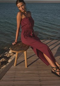 Bordeaux strapless kjole med indsnævret talje, frontknapdetaljer og sidesplid, parret med sorte spidse sandaler, siddende på en bænke af træ.
