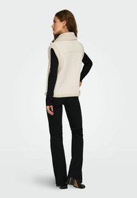 Creme farvet fleece vest med høj krave, der har en blød tekstur, båret over en sort langærmet skjorte, parret med sorte jeans og sko.