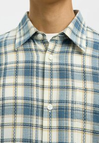 Gros plan sur une personne portant une chemise à carreaux bleue, blanche et beige par-dessus un t-shirt blanc, montrant le col et la partie supérieure de la poitrine.
