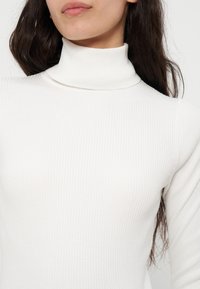 Femme portant un pull à col roulé côtelé blanc avec de longs cheveux foncés, montrée des épaules au menton sur un fond blanc.