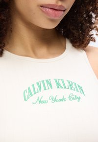 Κλείσιμο σε πρόσωπο με άσπρο ριμπ κορμάκι που γράφει πράσινα «Calvin Klein New York City», εμφανίζοντας τα χείλη και τα σγουρά μαλλιά.