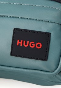 Geantă din material teal deschis, cu o suprafață texturată, având o bage black pe care este inscripționat "HUGO" în litere roșii îndrăznețe.