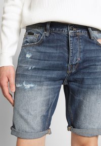 Denimshorts med en bleknad blå tvätt, uppvikta fållar, slitna detaljer och en femficksdesign. Har en liten logotyp nära midjebandet.