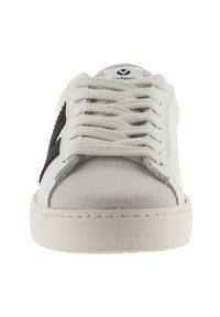 Vit sneaker med grå tåhuvud, tjocka vita snören, svart stjärndesign på sidan och beige sula sedd framifrån.