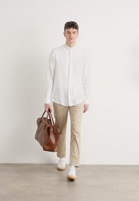 Witte linnen overhemd met knopen, beige chino's, bruine leren tas, witte sneakers met tan zolen, staand tegen een effen achtergrond.