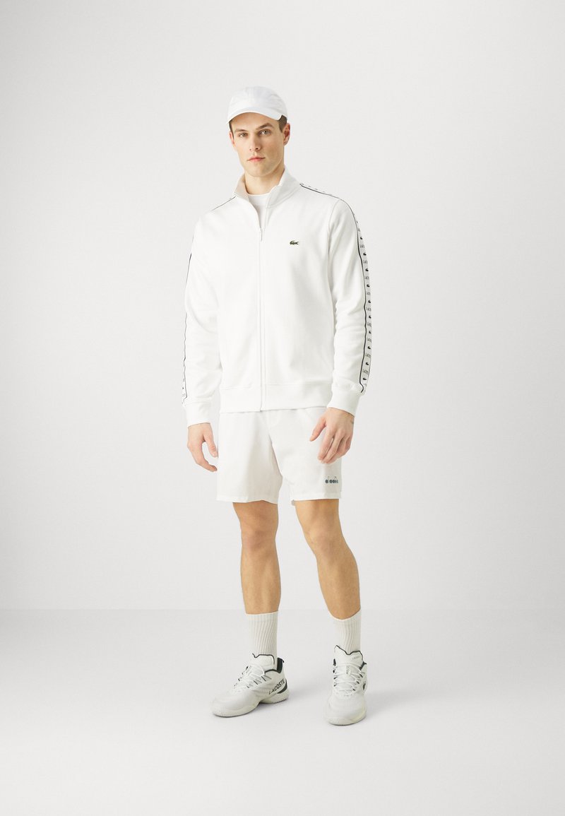 Witte sportieve tracksuit bestaande uit een zip-up jack en shorts, met zwarte accenten en logodetails, gecombineerd met witte sneakers en een pet.