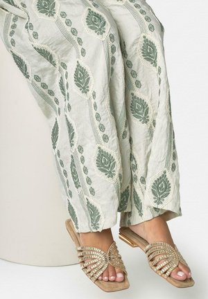 Pies con sandalias abiertas beige con adornos espejados, combinadas con pantalones verdes sueltos con estampado, sobre un fondo liso.