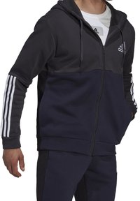 Sweat à capuche zippé bleu foncé avec une section supérieure plus claire, accents blancs à trois bandes sur les manches et poches avant. Fabriqué en tissu doux.