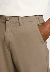 Homme portant un pantalon beige avec un bouton et une fermeture à glissière, main dans la poche, et une chemise blanche rentrée visible à la taille.