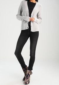 Svetle šedý pletený cardigan s vroubkovanou strukturou a předními knoflíky, v kombinaci s černými skinny džíny a kotníkovými botami s květinovým vzorem.